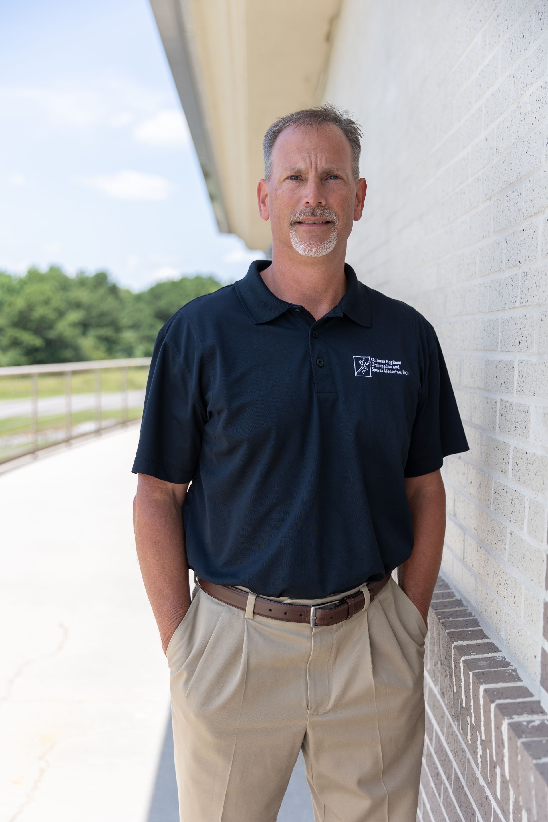 Tommy Mullican, PA-C in a blue shirt is leaning against a brick wall with his arms crossed.
