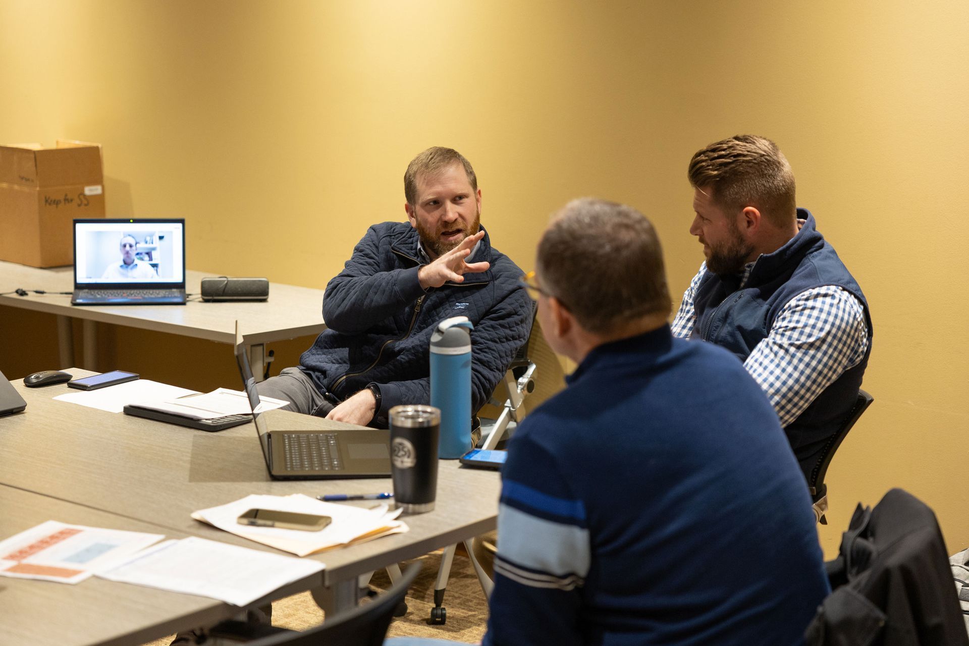Three men in a meeting, one gesturing while others listen at a table.