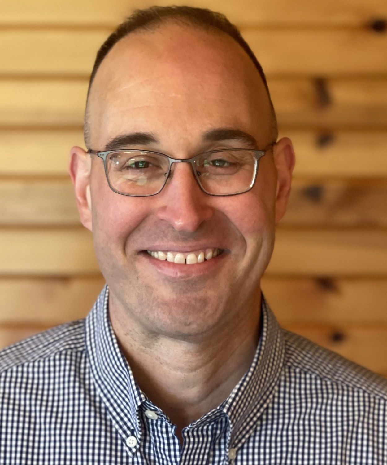 Smiling man with glasses, wearing a blue and white checkered shirt, in front of a wooden background.