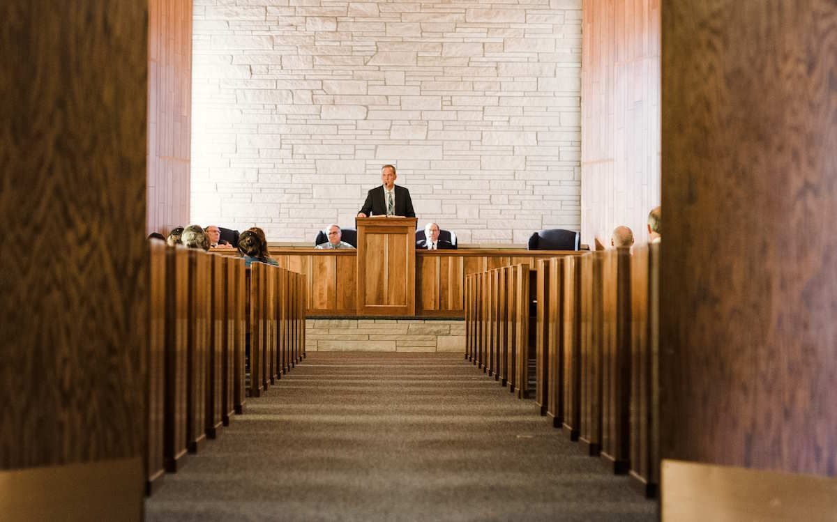 A man speaks from a wooden pulpit in a church, viewed from the aisle between pews. Light stone wall backdrop.