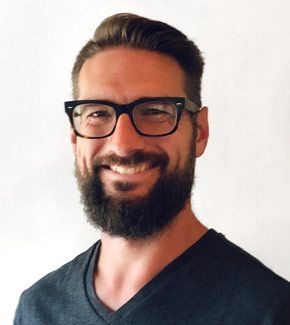 Man with a full beard and glasses smiles, wearing a dark v-neck shirt, against a white background.
