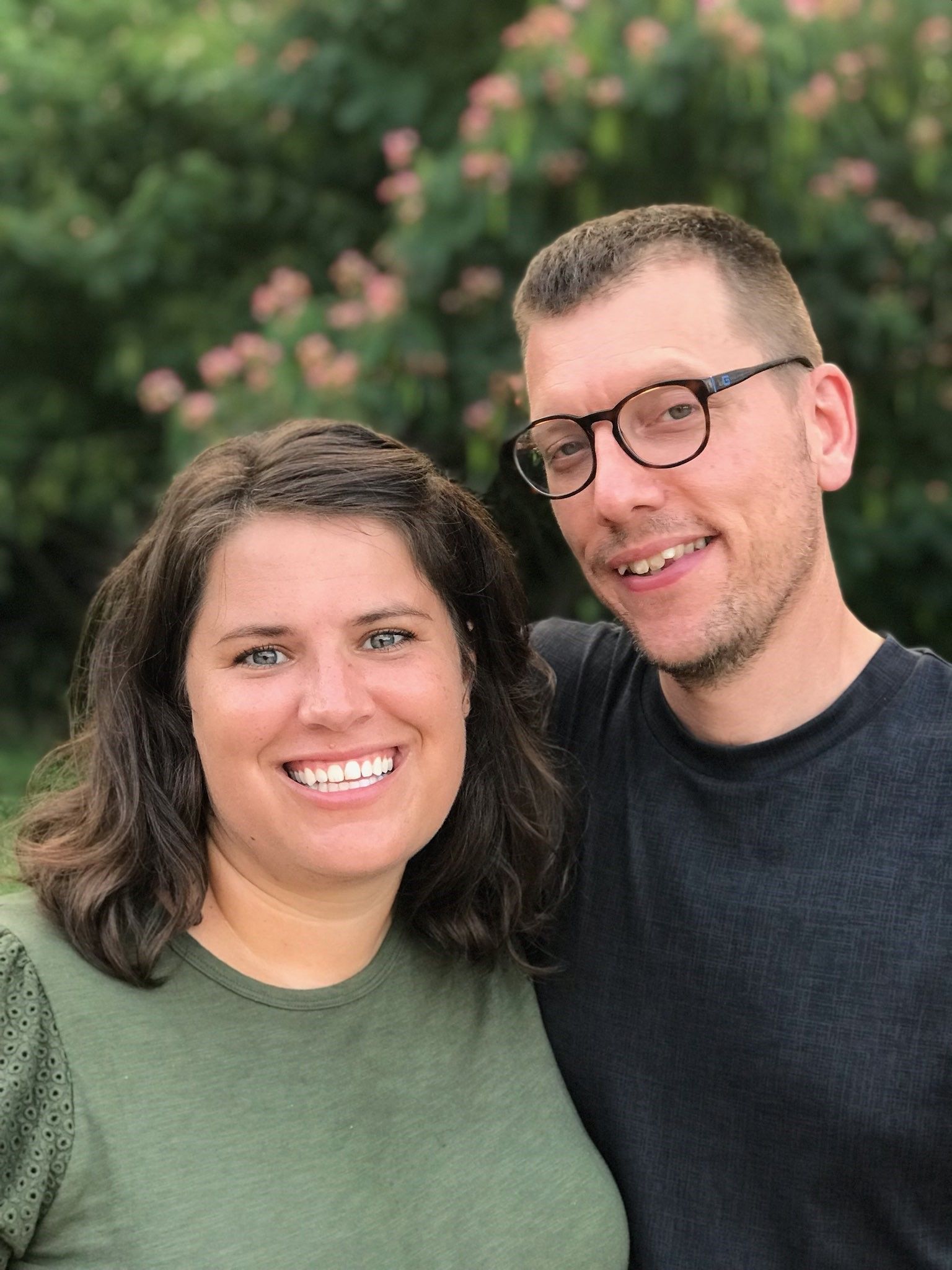 Smiling couple, a man with glasses and a woman in a green shirt, pose outdoors. Blurred green foliage in background.
