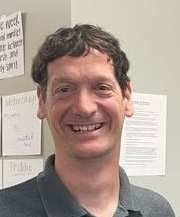 A smiling man with short brown hair, wearing a gray collared shirt, standing against a wall with posted papers.