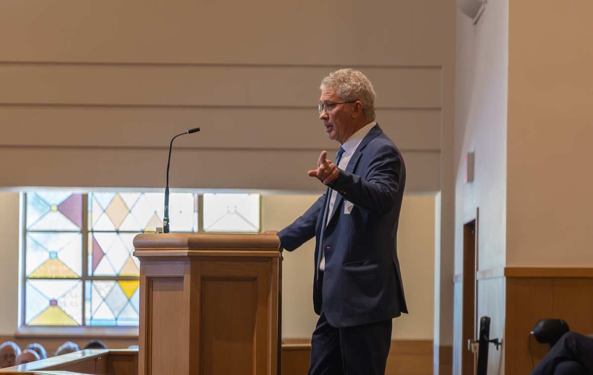 A man in a suit gestures while speaking at a podium in a wood-paneled room with stained glass window.