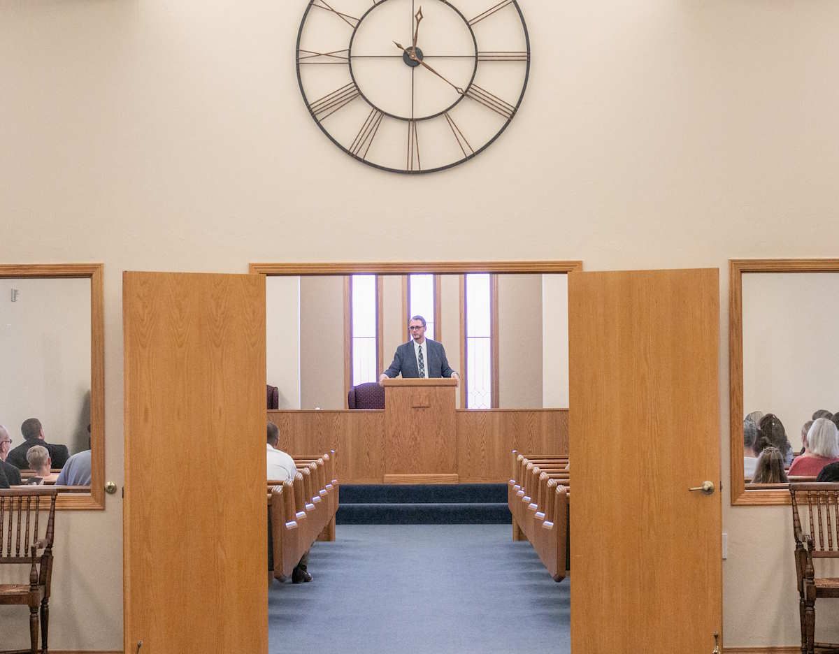 A person speaking from a wooden podium inside a church. View through open doors, with pews and a clock visible.