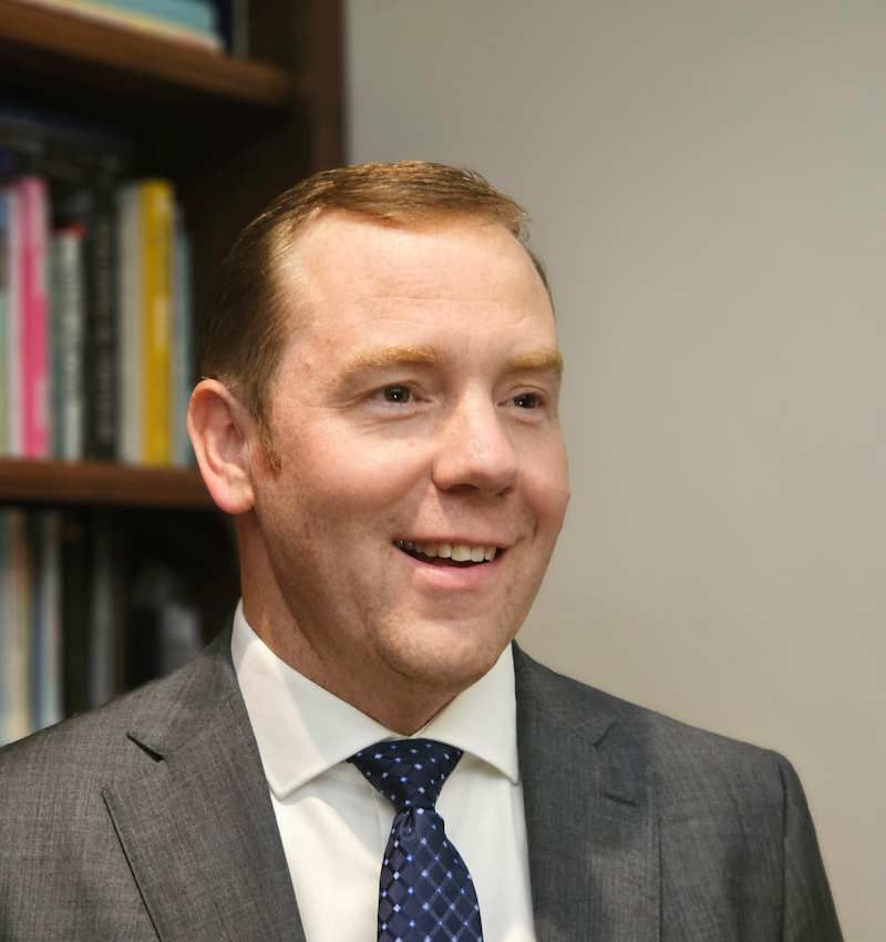 A man in a suit smiles broadly, with a bookshelf in the background.