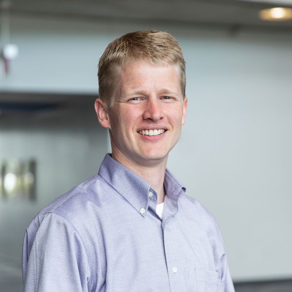 A smiling fair-skinned man with blonde hair wears a light blue button-down shirt in a modern building.
