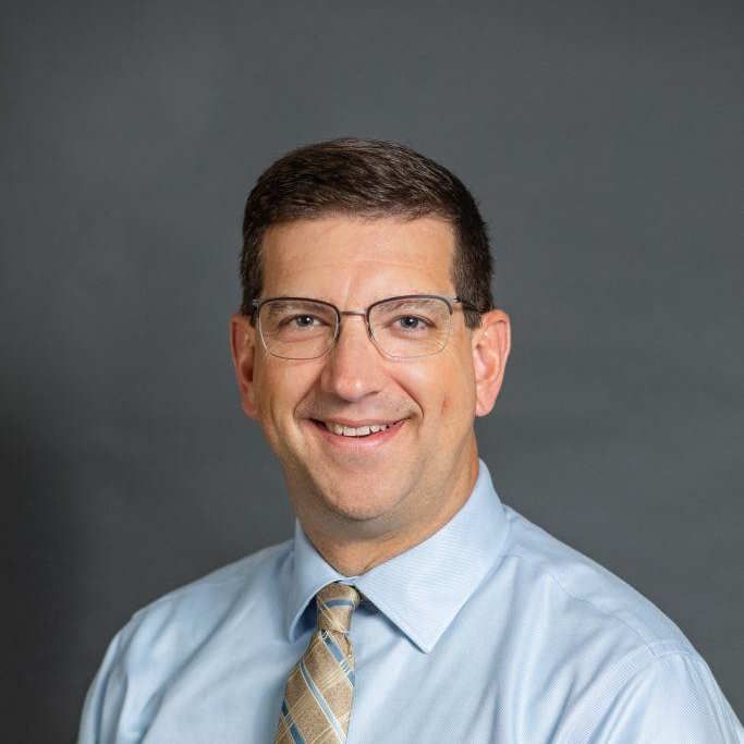 Smiling man with glasses, wearing a light blue shirt and patterned tie against a gray backdrop.