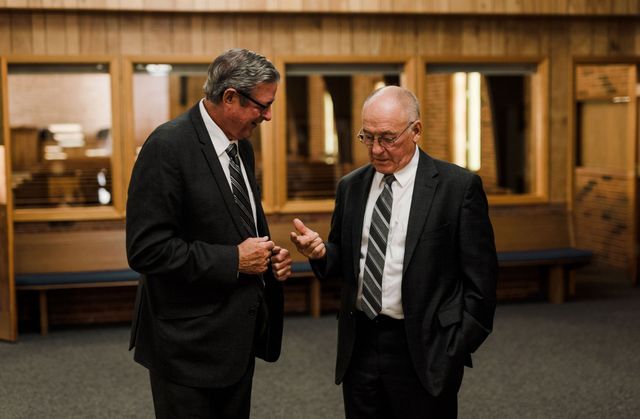 Two men in suits conversing indoors. One gestures while the other listens intently.