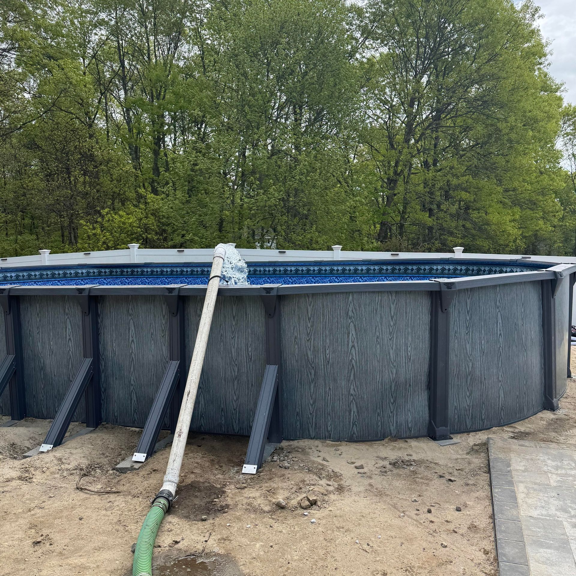 Above-ground pool with gray wood-like siding being cleaned by a person with a long pole and hose. Trees in the background.
