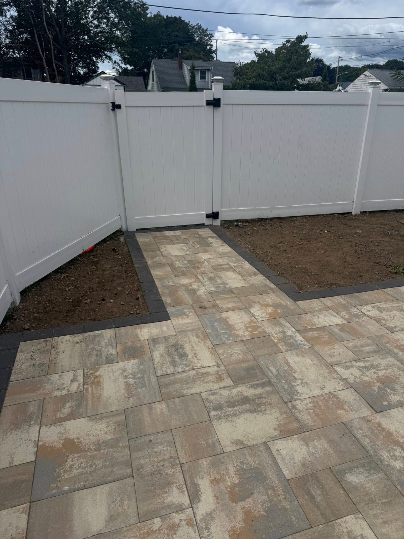 White vinyl fence with a gate, leading to a paved patio area. Dirt borders the walkway.