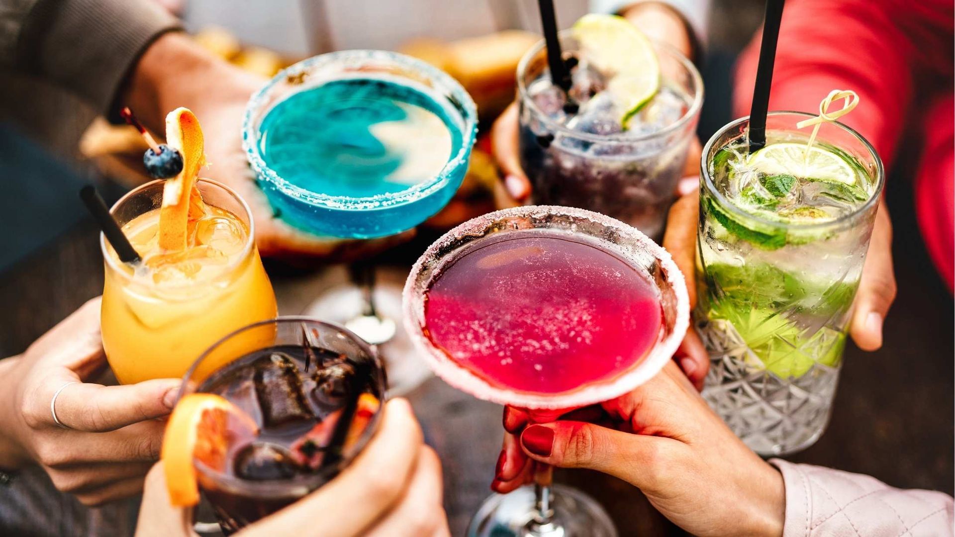 Hands clink colorful cocktails together at an outdoor gathering. Drinks include blue, purple, green, and orange hues.