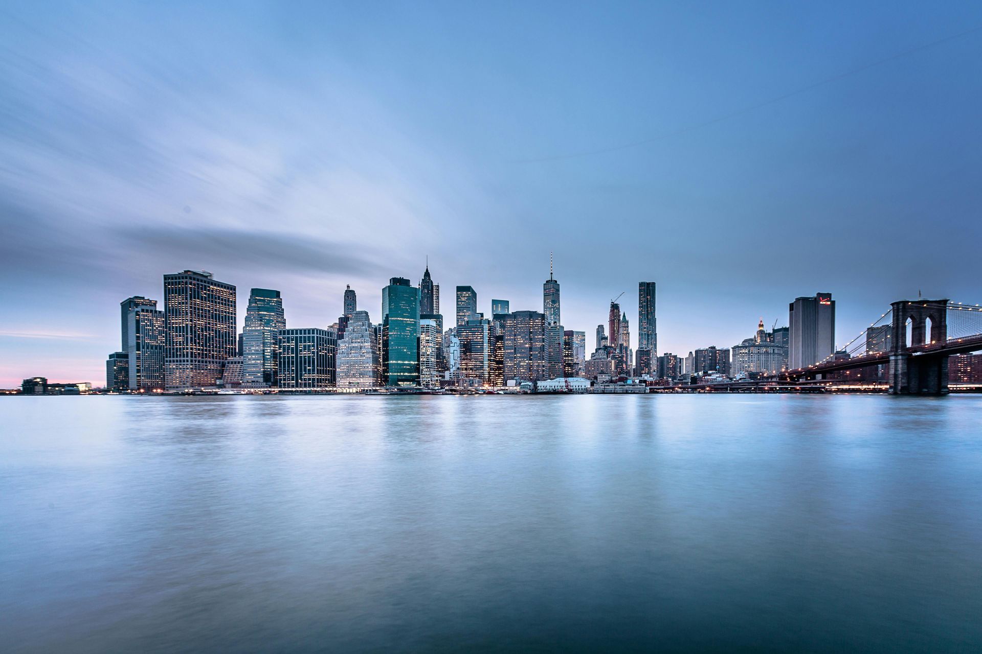 New York City skyline at dusk, buildings reflecting in the water. Brooklyn Bridge on the right.