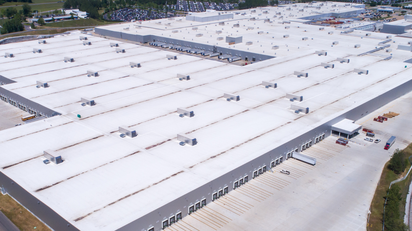 Aerial view of a large white industrial warehouse complex with parking lots and surrounding roads.