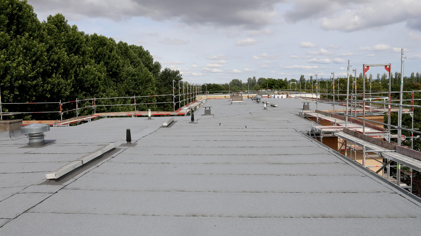 Rooftop under construction with gray roofing, scaffolding, and trees in the background