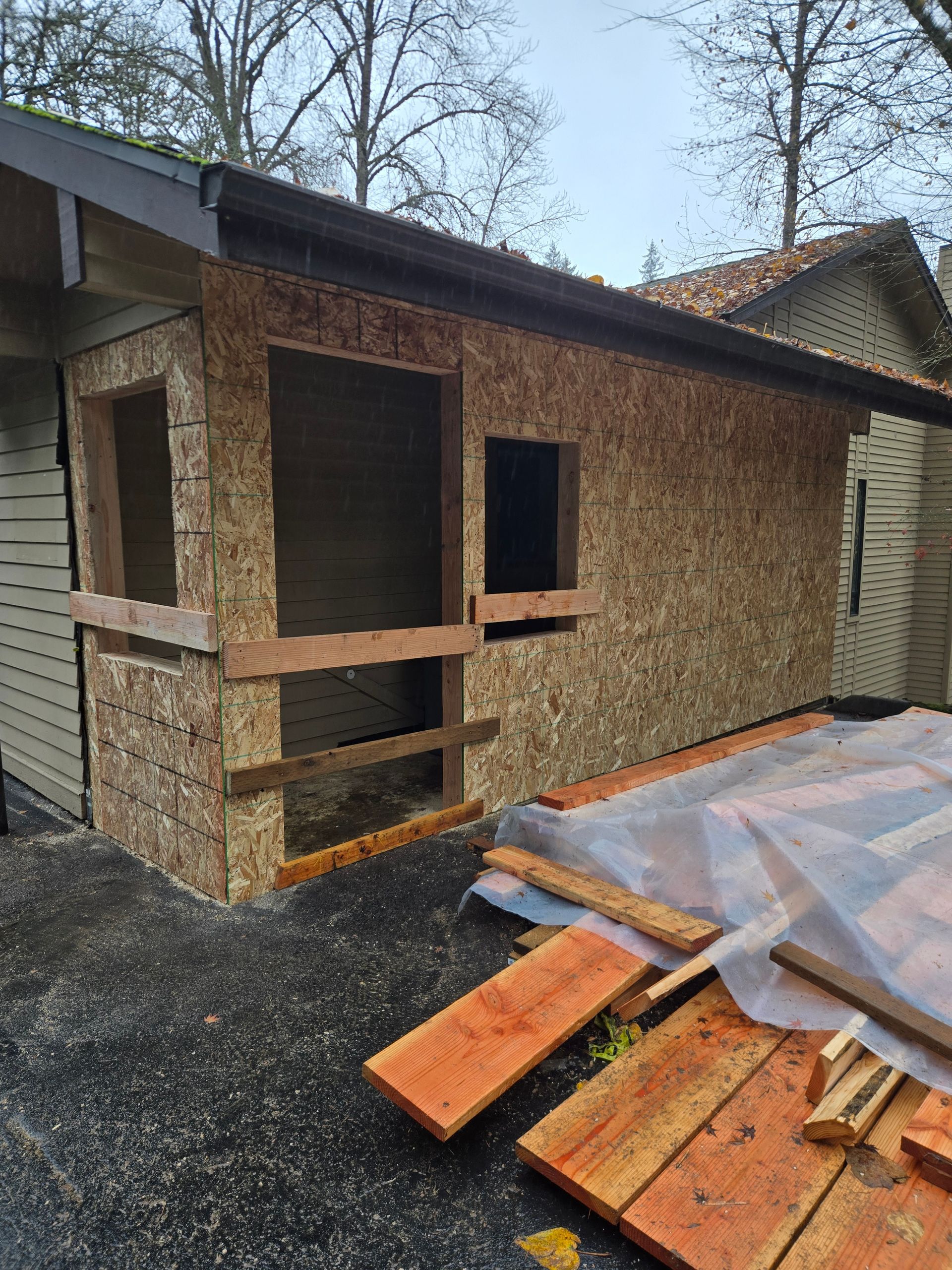 Partially renovated small house with exposed wood siding, window openings, and lumber stacked outside