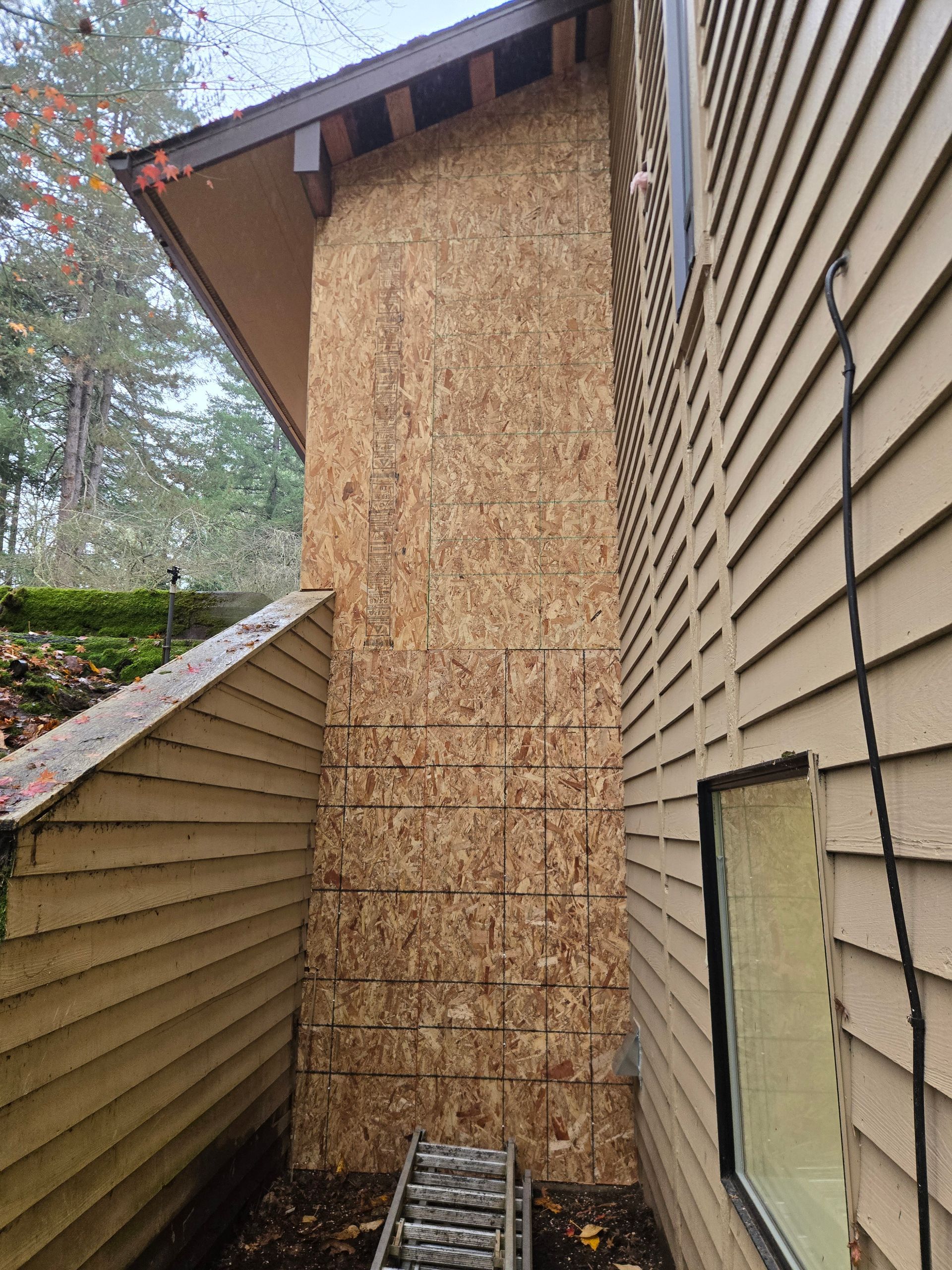 Narrow exterior side yard with a plywood-covered wall, beige siding, and a steep roof edge after storm damage.