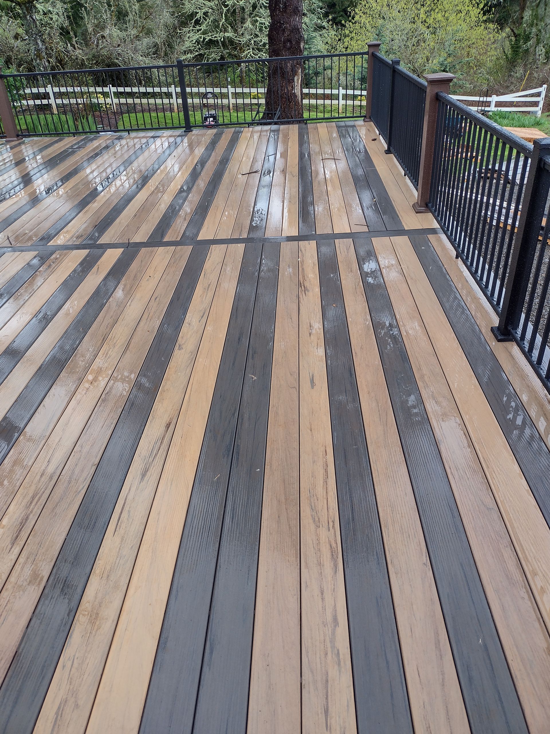 Wooden deck with alternating gray and tan planks beside a black metal railing and trees beyond
