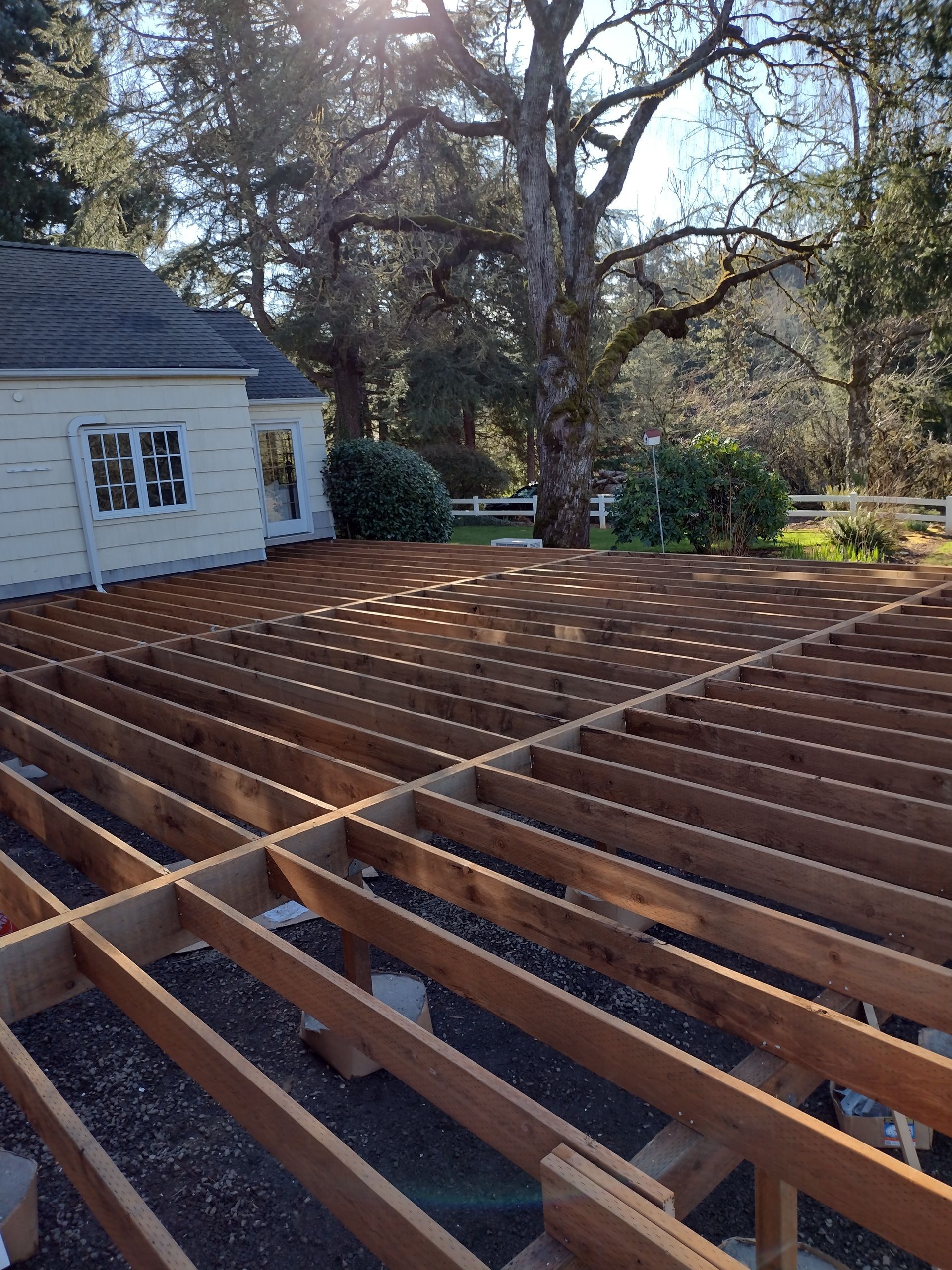 Wooden deck framing under construction beside a white house and large tree in a backyard
