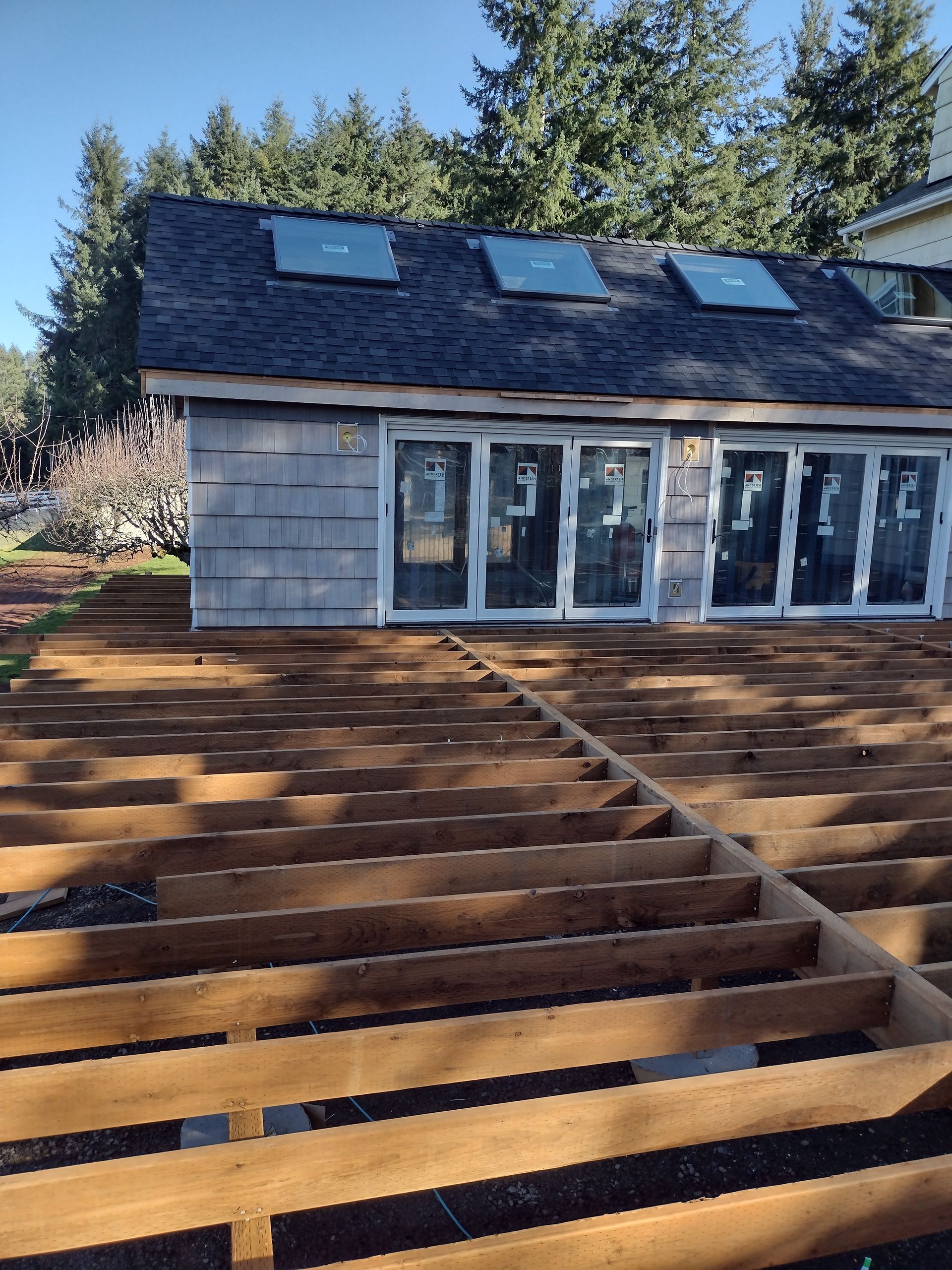 Backyard patio under construction with wooden deck framing and a house with skylights in the background