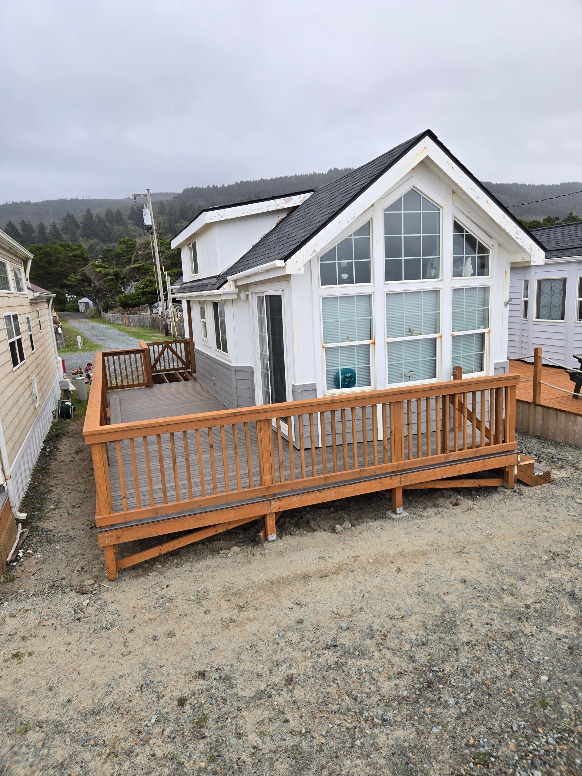 White beach house with large front deck and sloped roof on a gravel lot near trees and neighboring homes