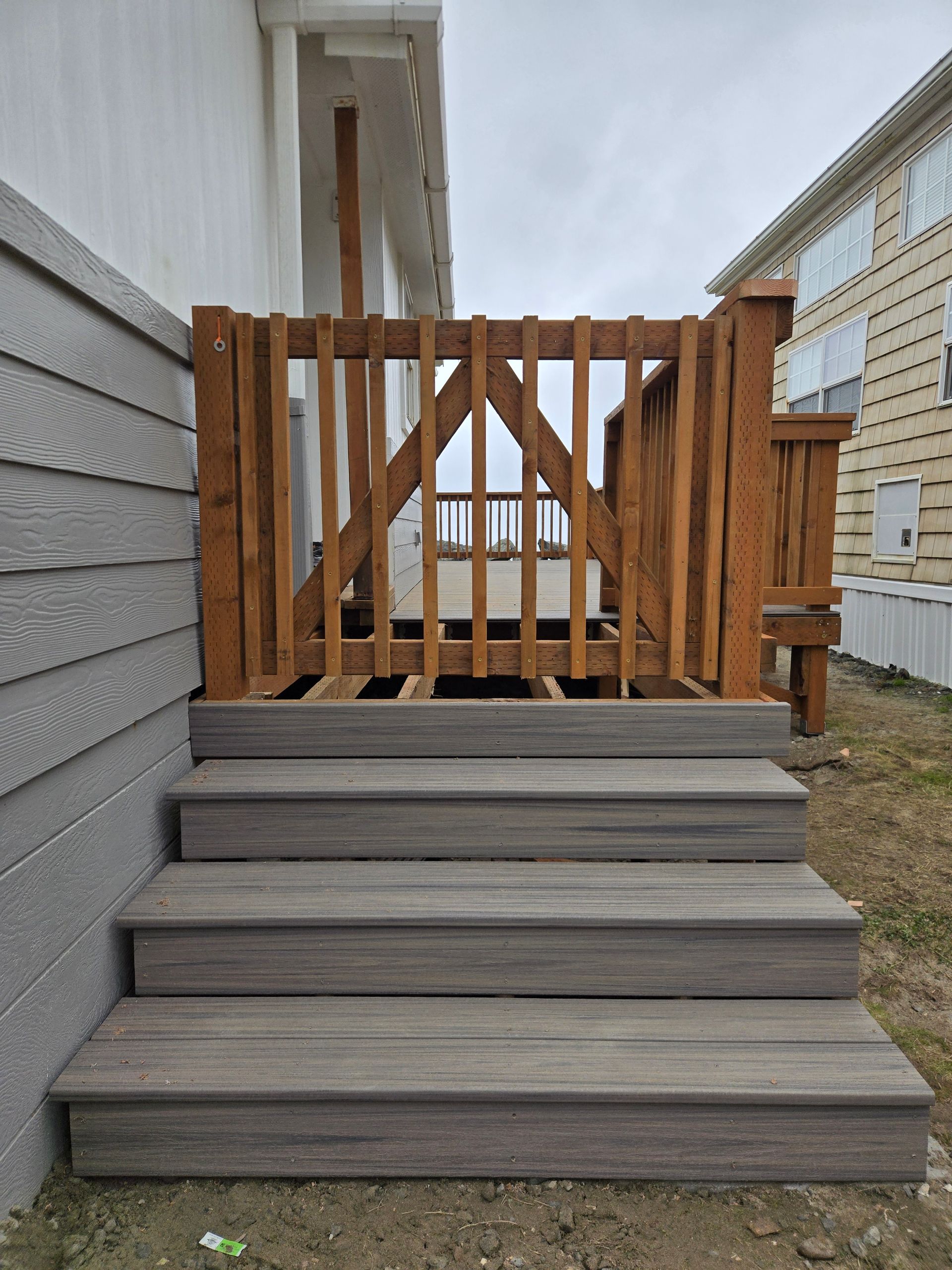 Wooden front steps leading to a small deck with a tan slatted railing beside a house.