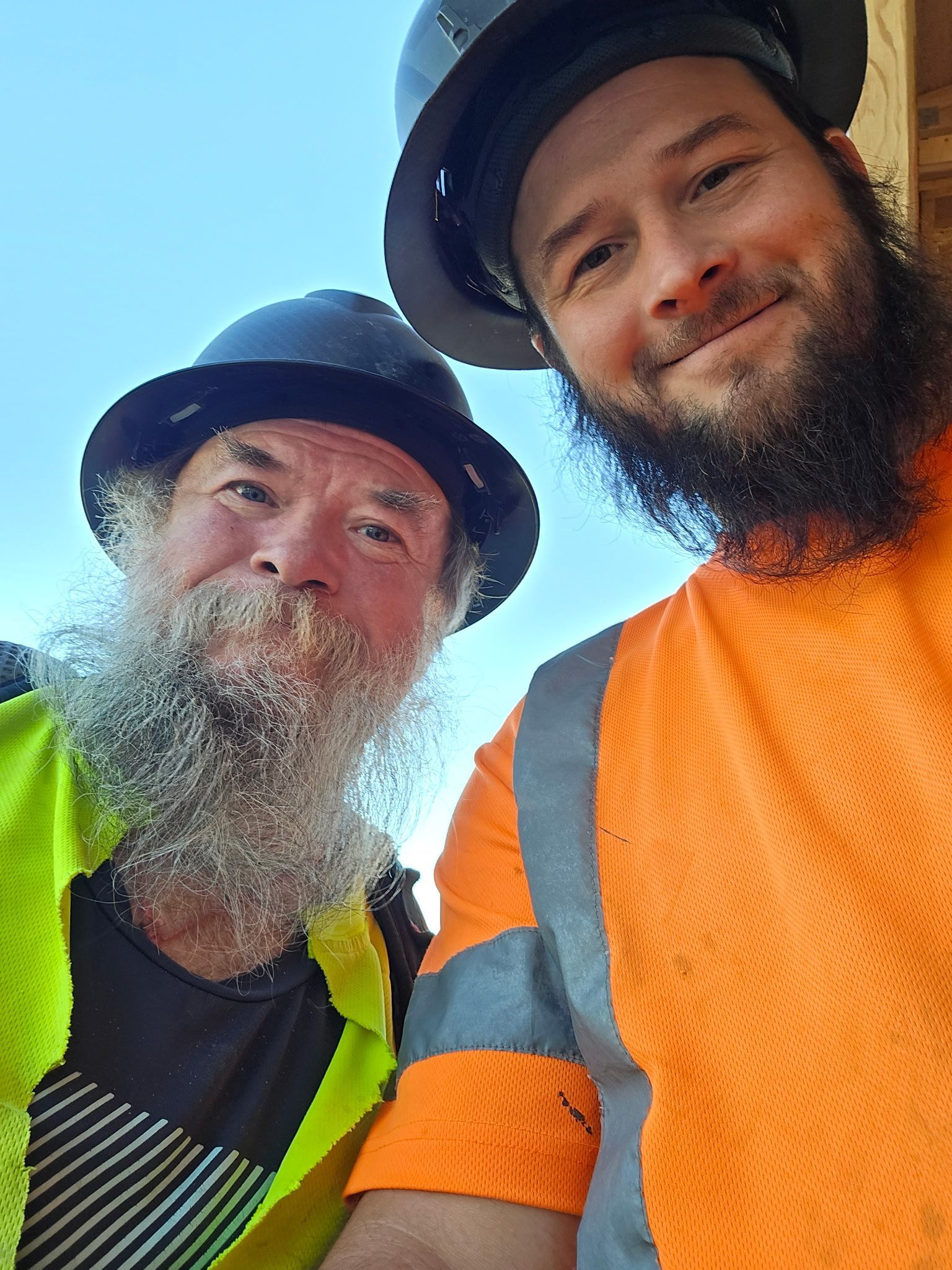 Two workers in hard hats and hi-vis shirts smile at the camera outdoors