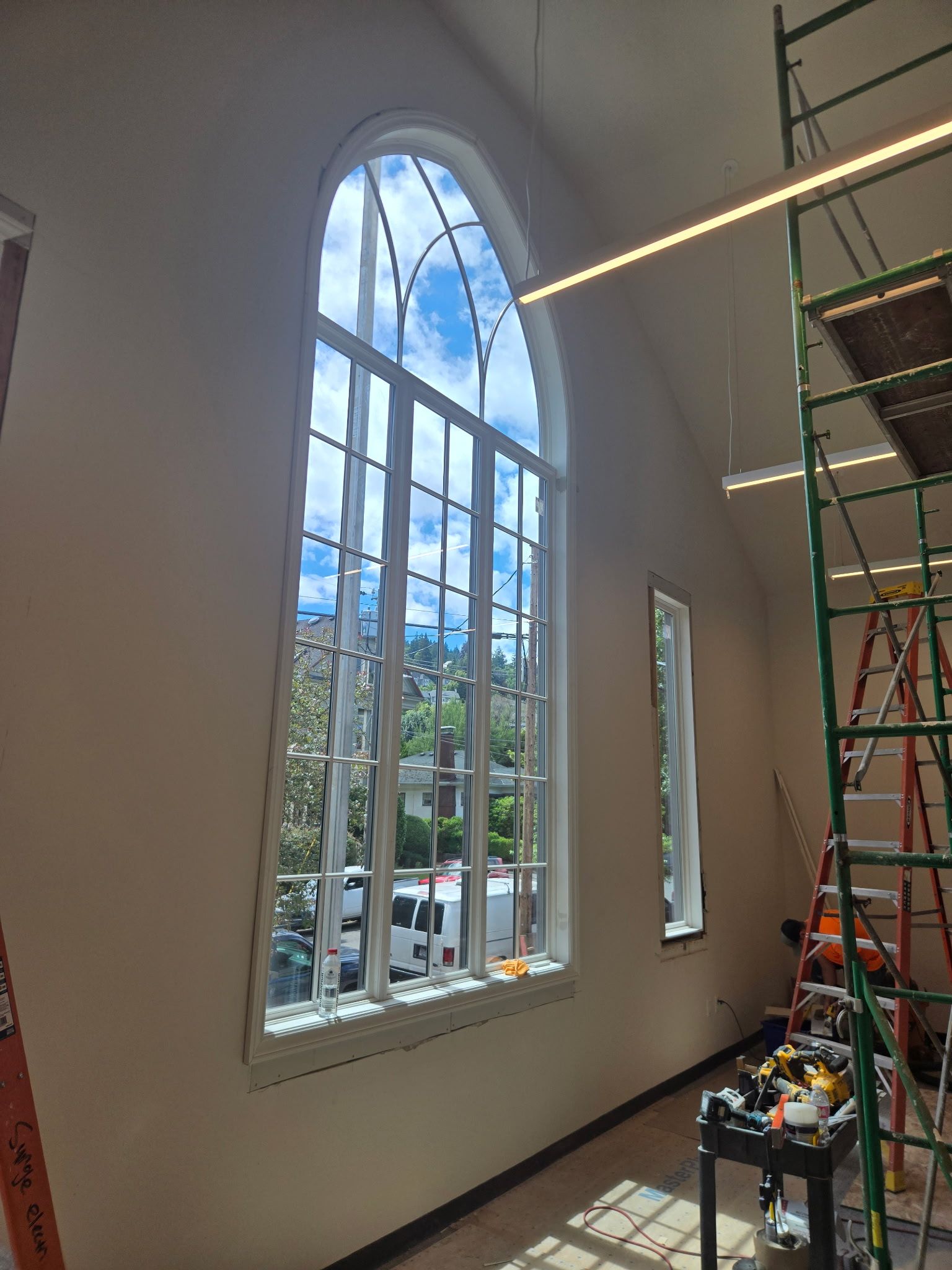Tall arched stained-glass window in a white interior, with scaffolding and construction tools nearby.