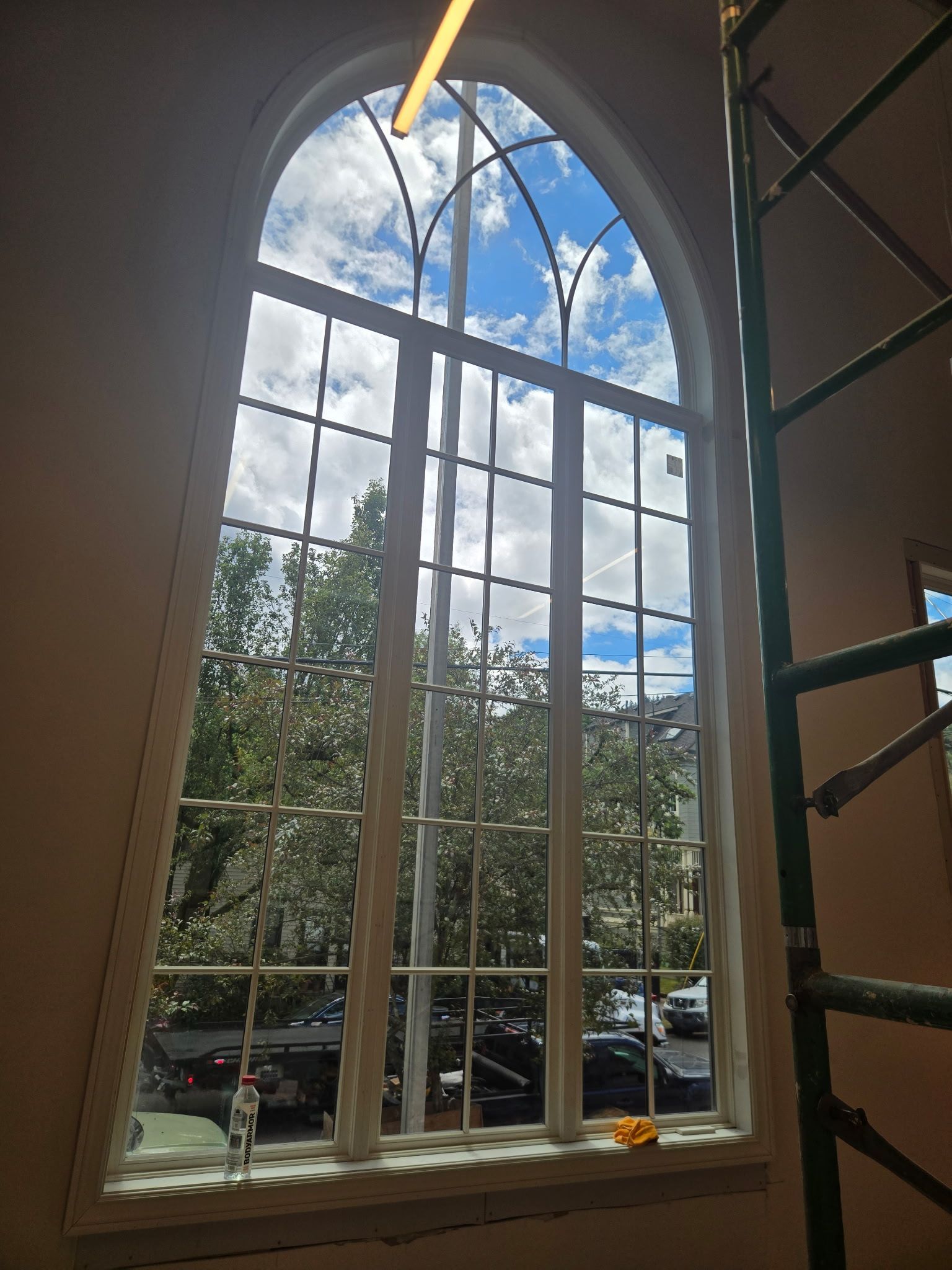 Arched window with white mullions, trees outside, and a yellow ladder inside near the right side.