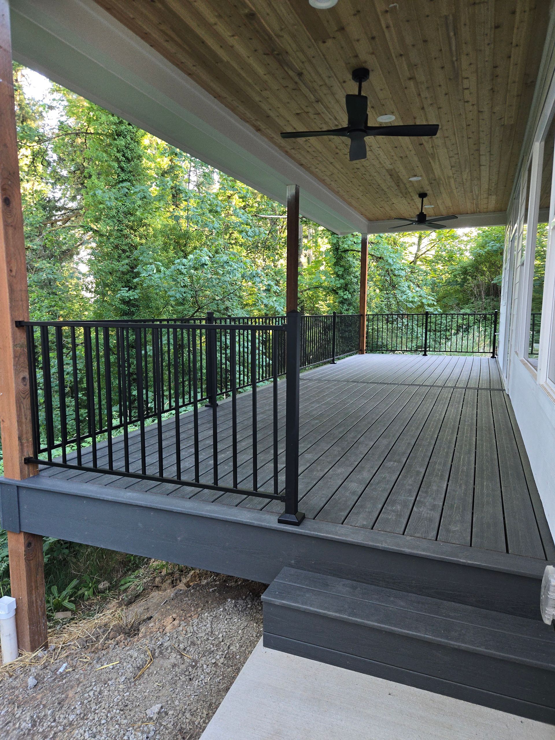 Covered wooden porch with black railings, steps, and a ceiling fan overlooking trees and gravel below