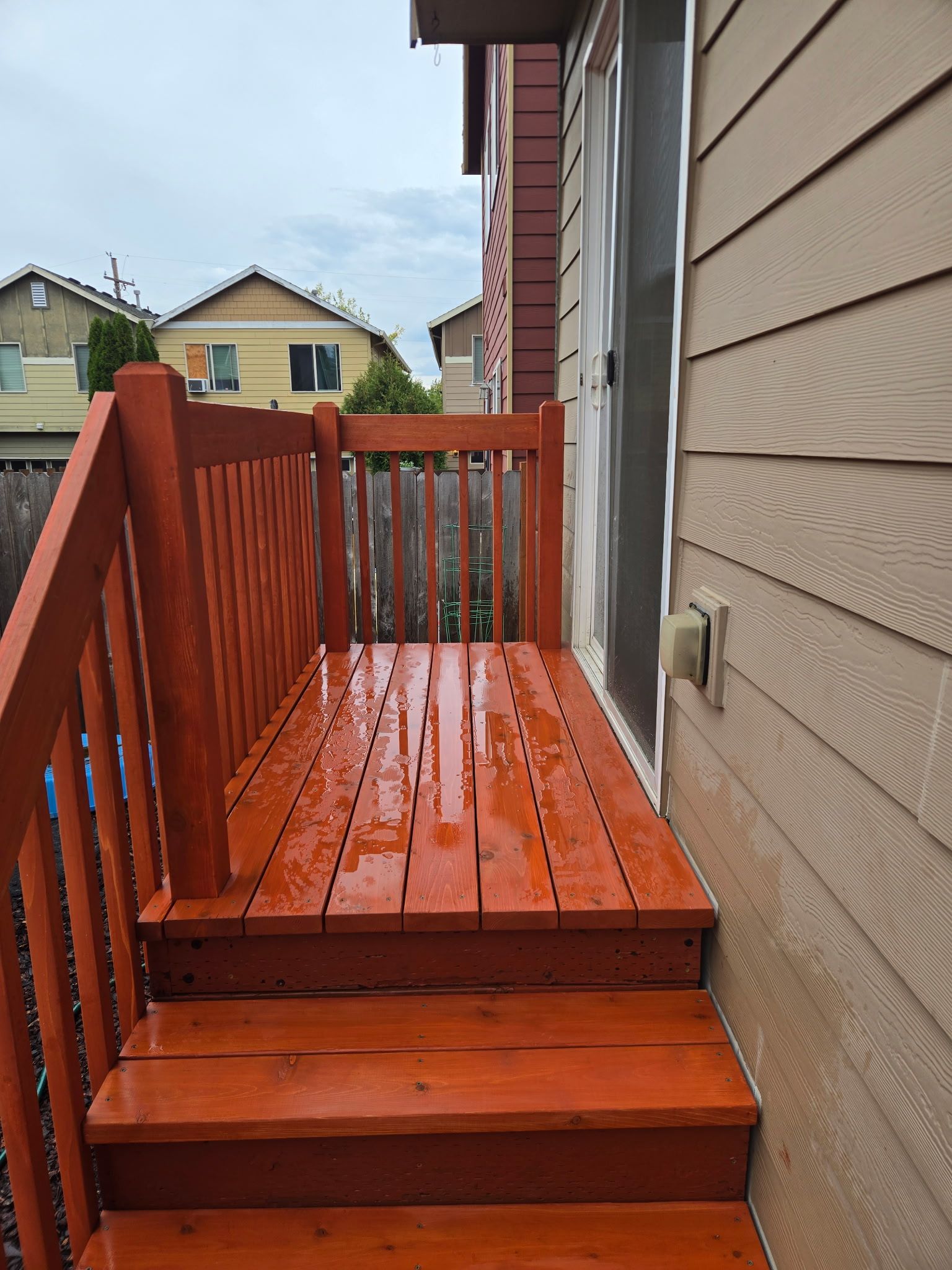 Wet orange wooden deck stairs beside a beige house, leading to a small balcony.