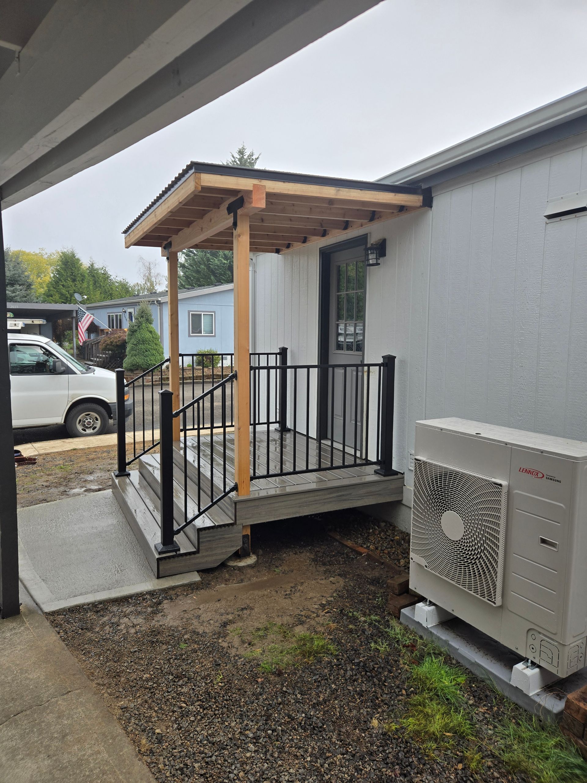 Small covered porch with black railing beside a gray building and outdoor AC unit.
