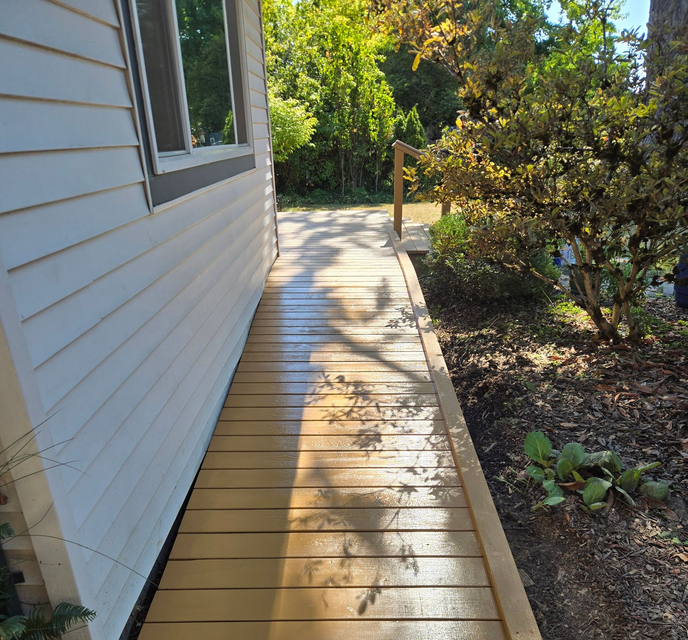 Wooden side walkway beside a white house, with a bush-lined garden and sunlight casting shadows.