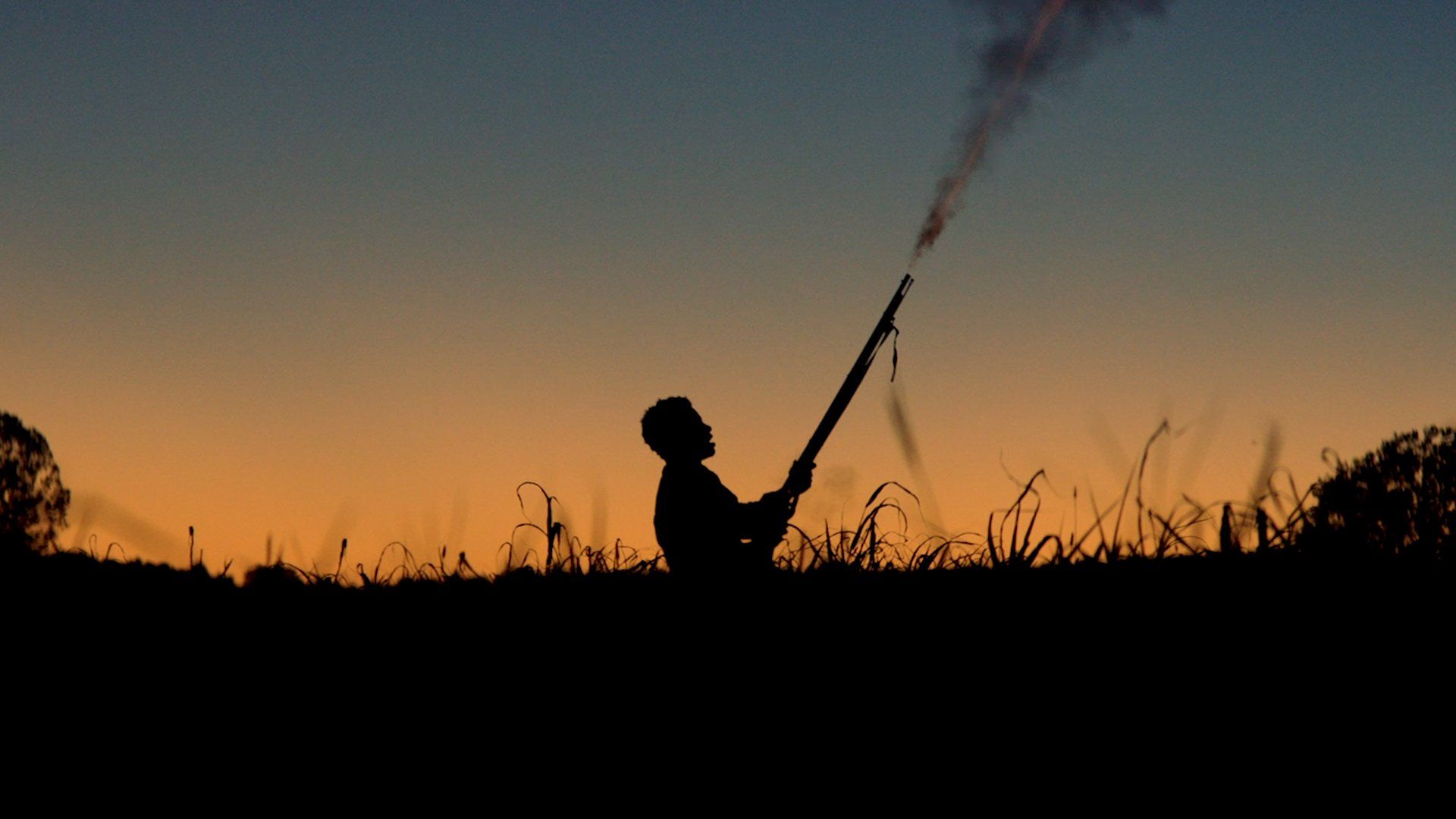 Silhouette of a person launching a skyrocket at dusk in a grassy field
