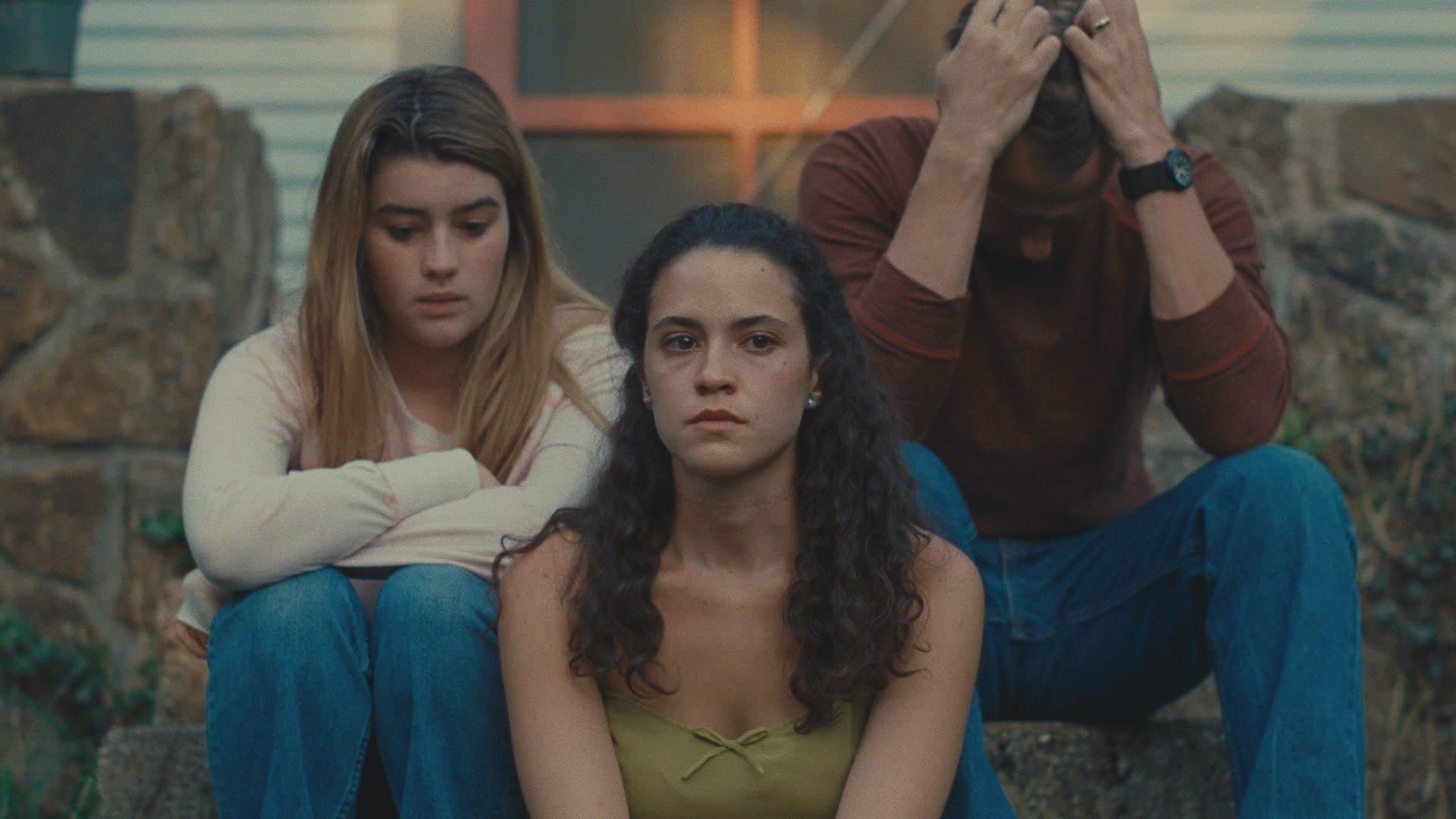 Three people sit on stone steps, looking somber and contemplative.