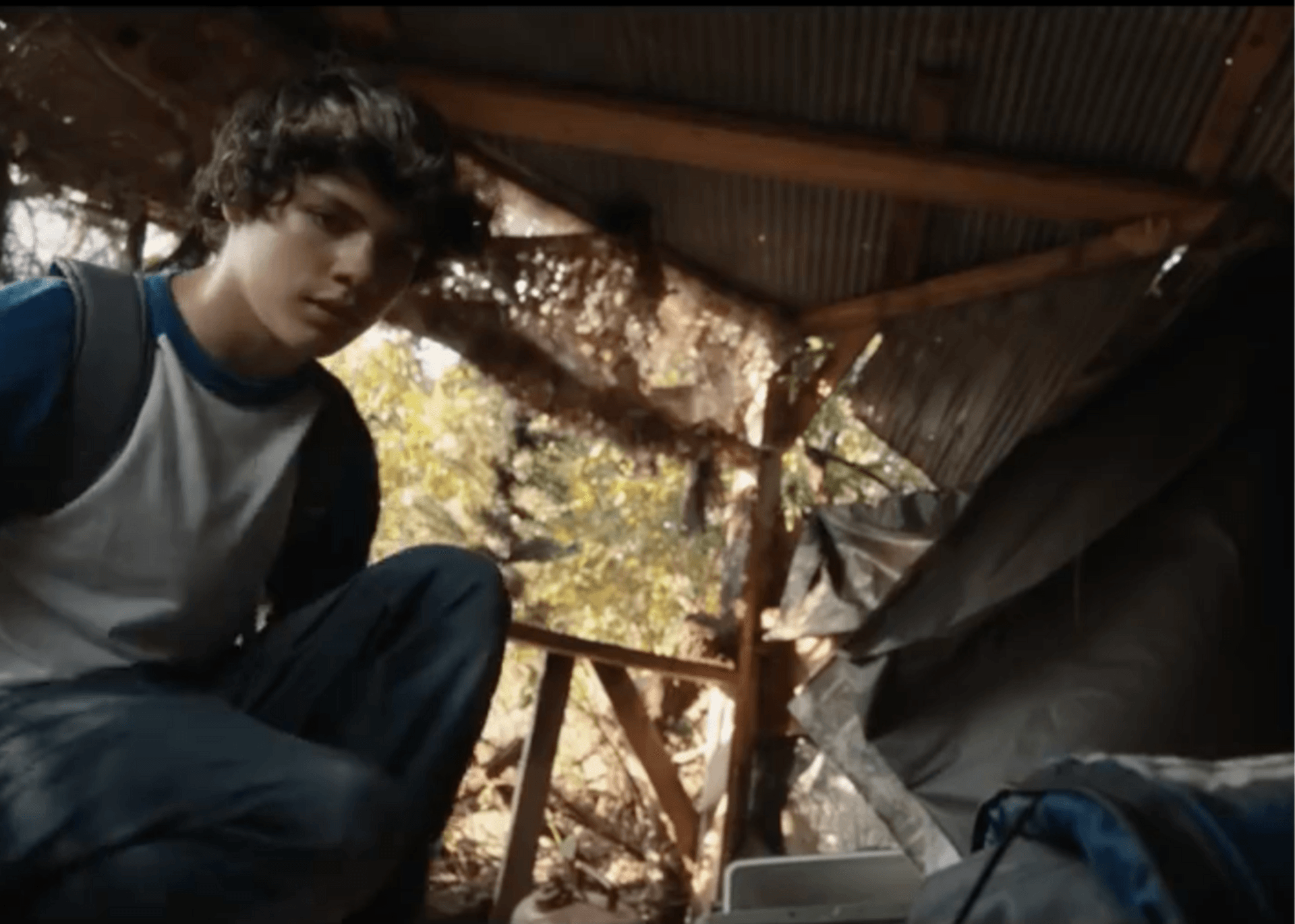 Person crouching inside a rustic shelter with a corrugated metal roof, looking toward the camera.