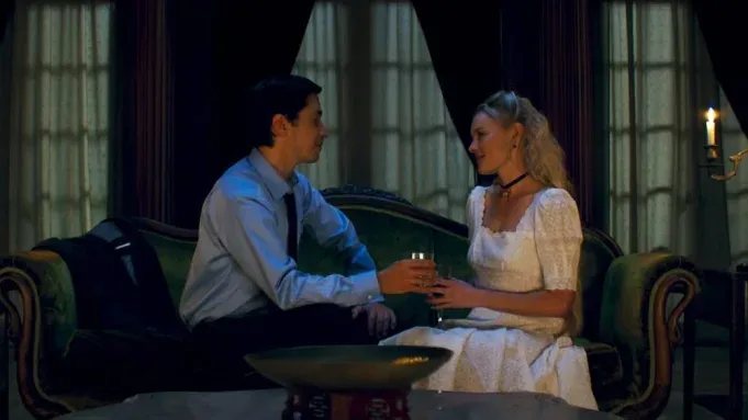 Two people sit facing each other in a dimly lit room, holding hands and talking at a small table.