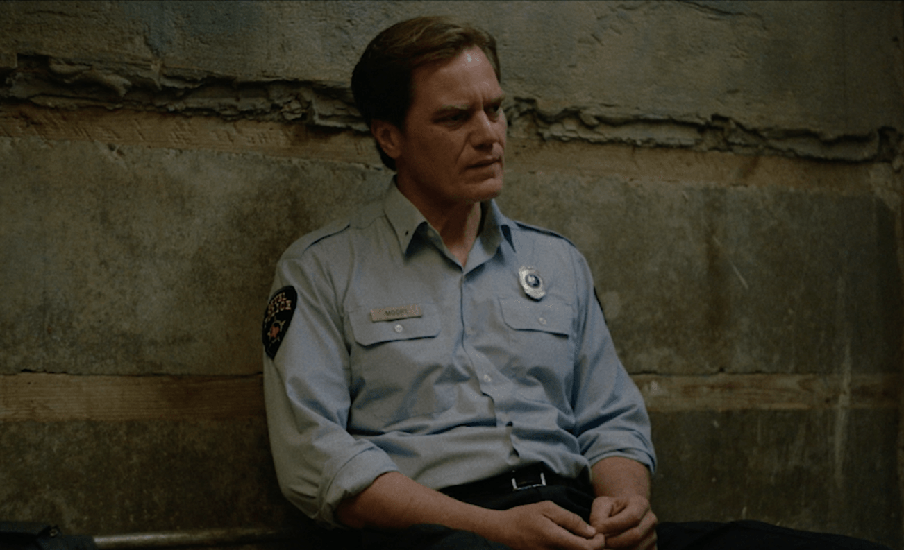 Man in a light blue uniform shirt sits against a weathered stone wall, looking down solemnly.