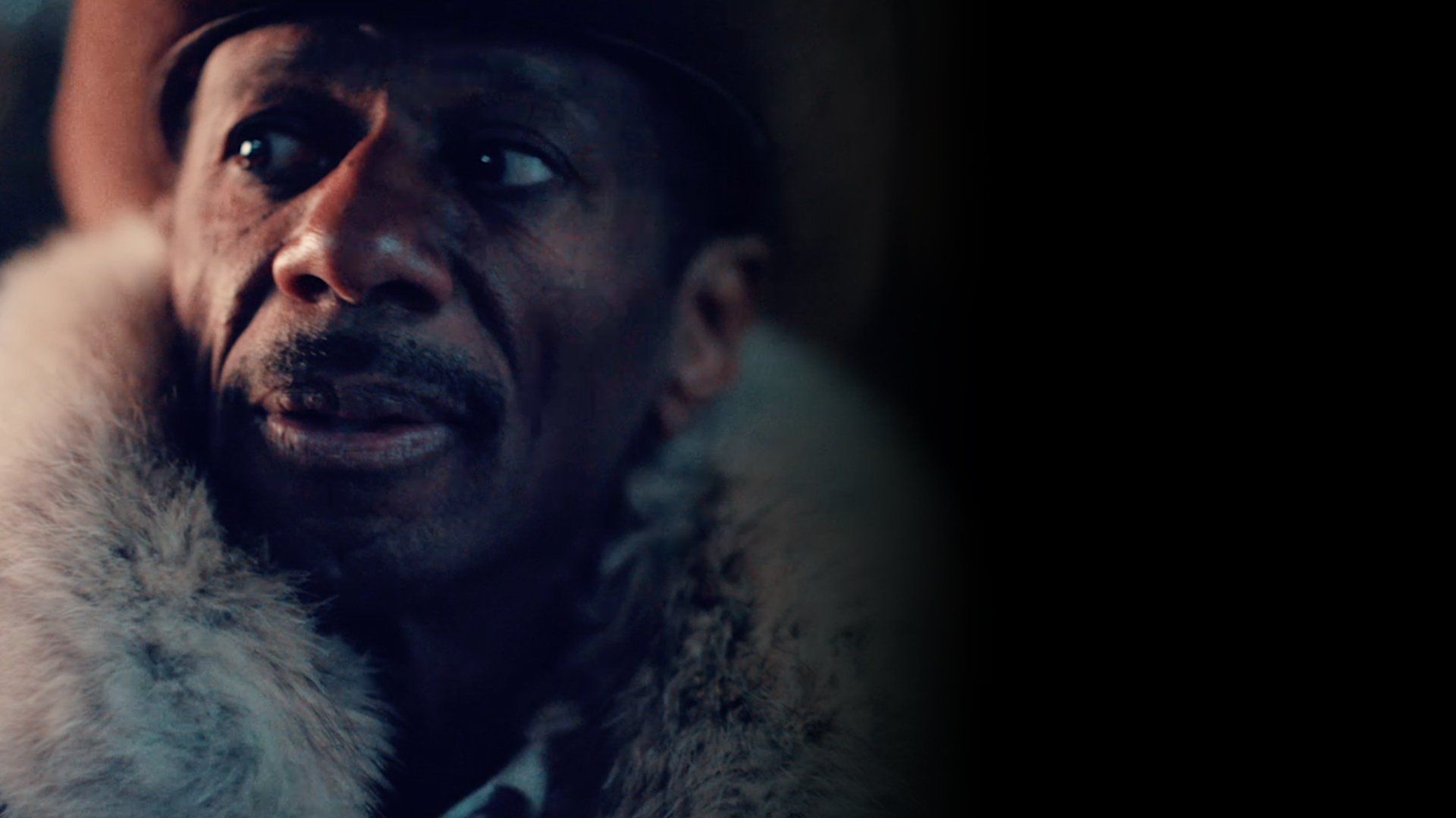 Close-up of a person in a fur-collared coat and hat, looking off to the side in dim light.