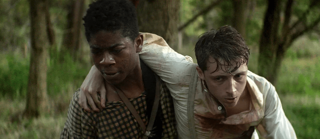 Two men struggle through a muddy forest, one carrying the other on his back.