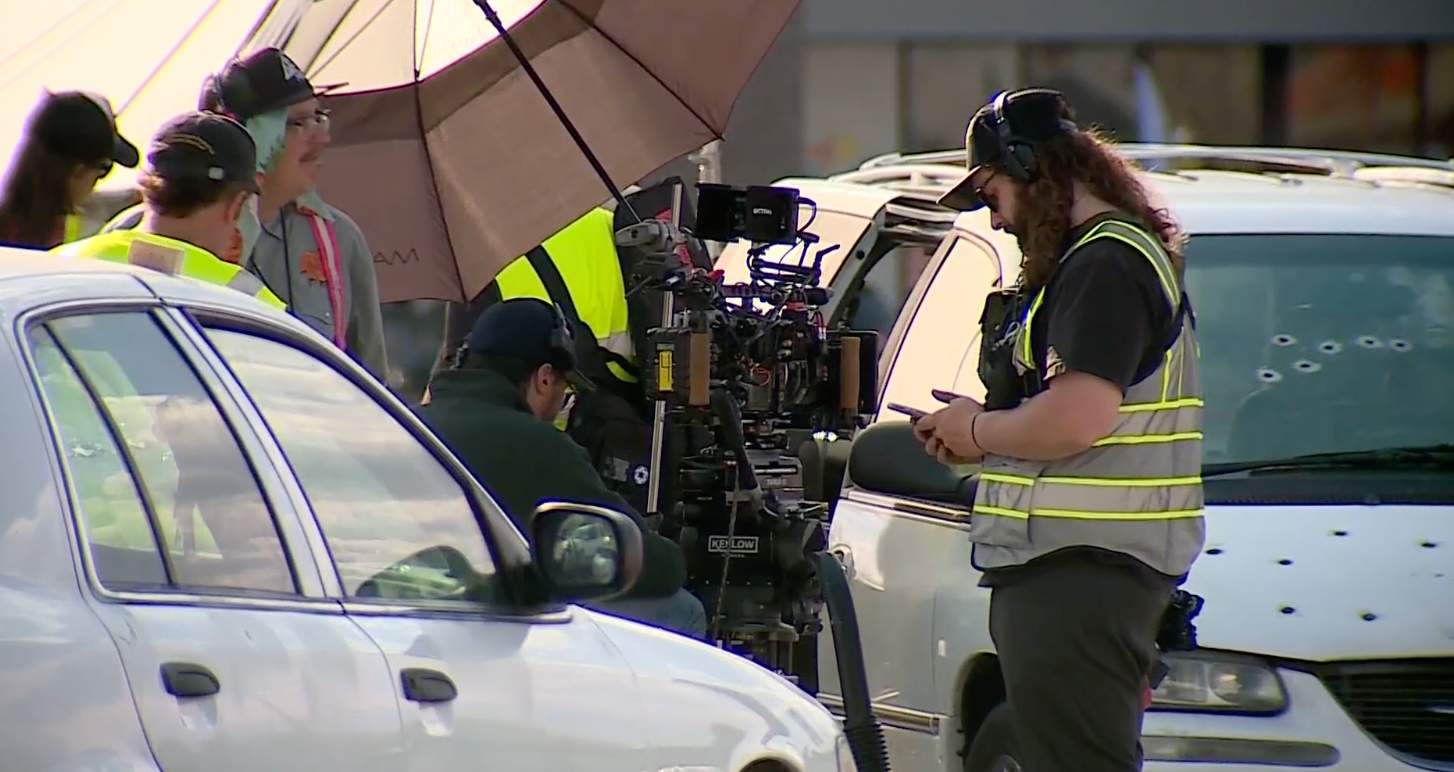 Film crew filming beside parked cars, with cameras and equipment under a large umbrella.