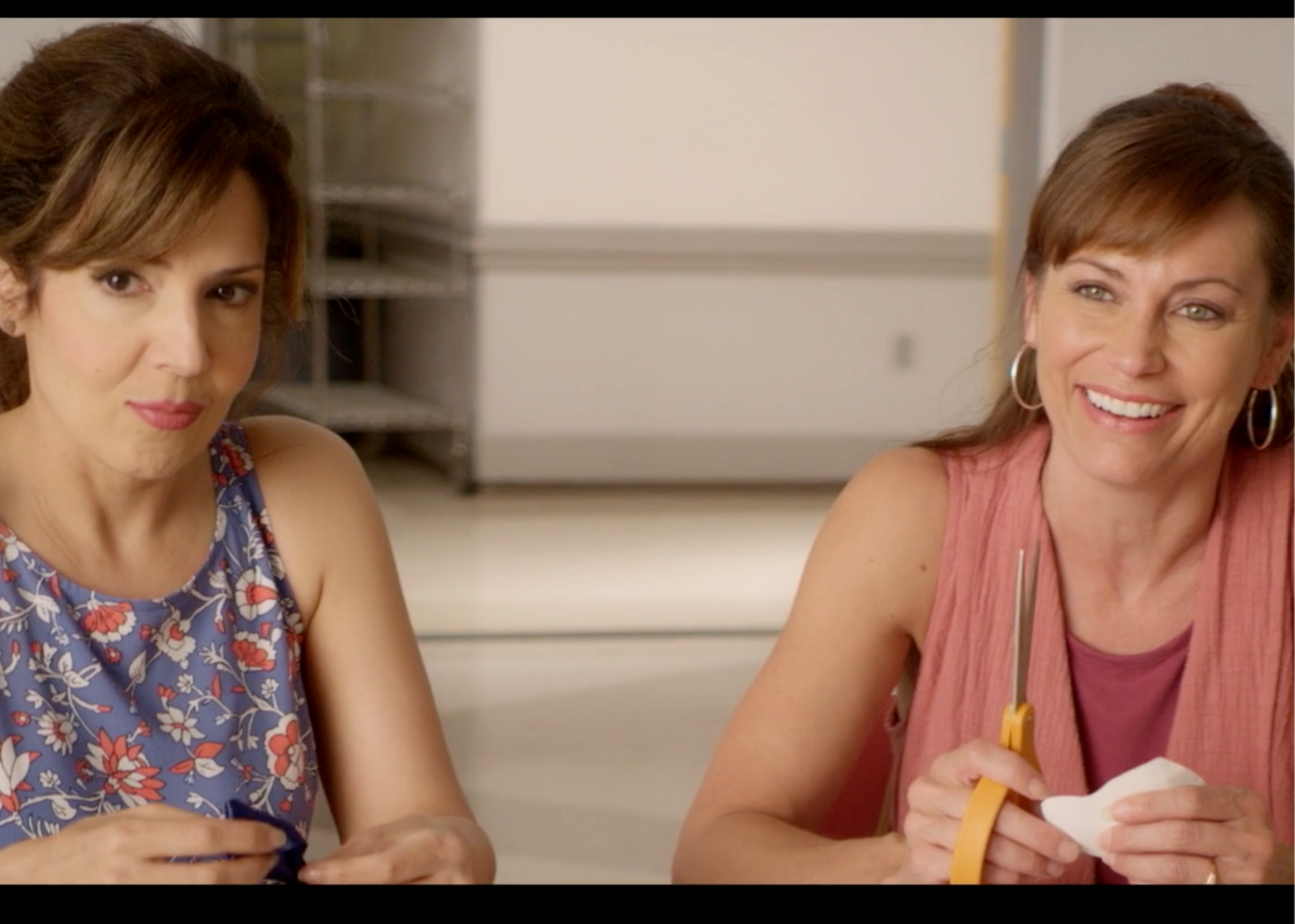 Two women sitting at a table, smiling and talking while holding pens and paper in a bright room