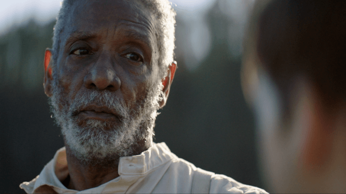 Close-up of an older man with a gray beard, looking solemn outdoors in warm light