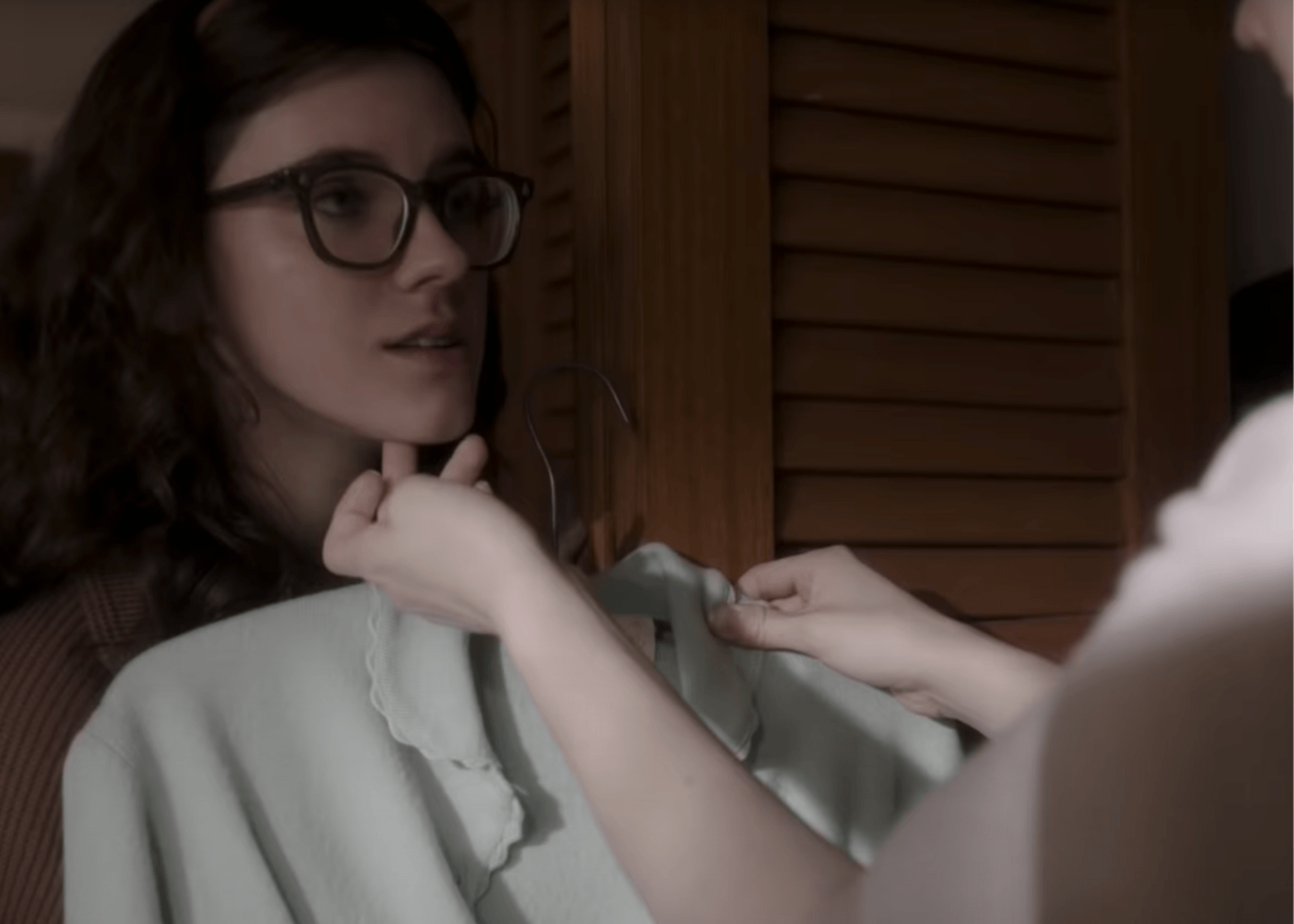 Woman adjusting a light green top while seated indoors, with a hand near her chin and glasses on.