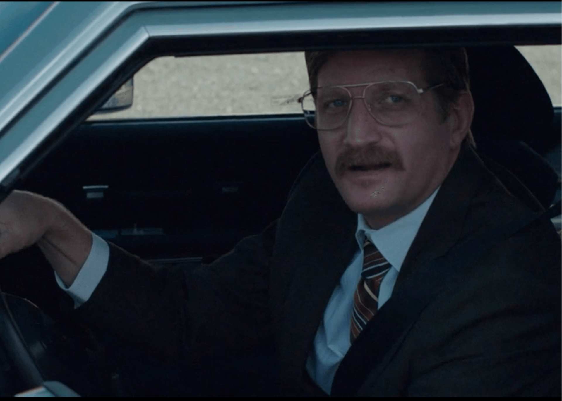 Man in a suit and glasses sitting in a car, looking toward the camera.