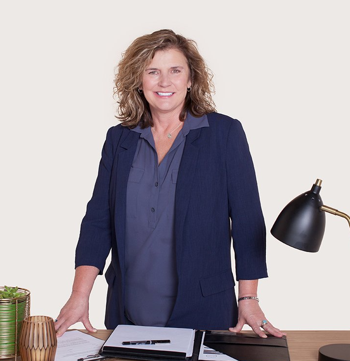 Woman standing at a desk in an office, wearing a navy blazer beside a black desk lamp and notebook.