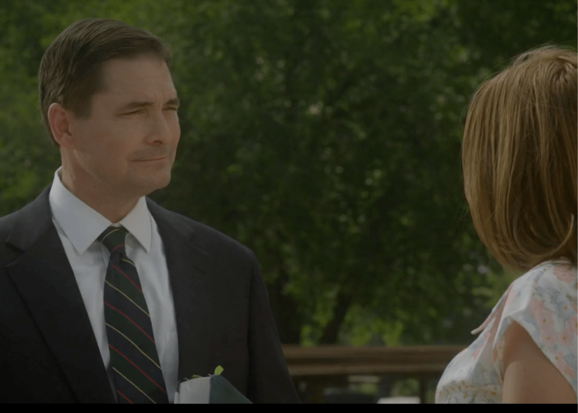 Man in dark suit talking to a woman outdoors, with trees in the background.