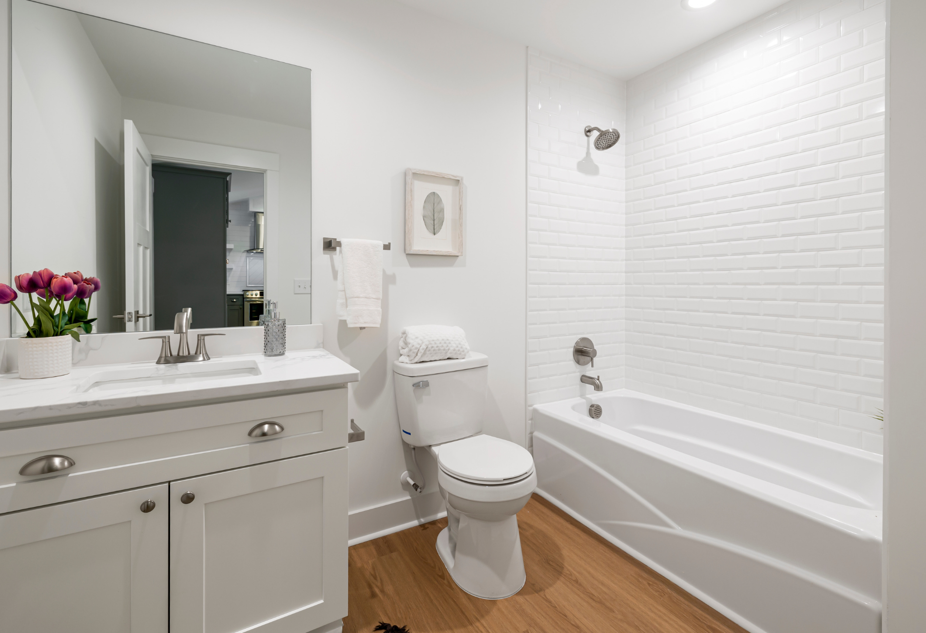 White bathroom with a vanity, toilet, and bathtub; light wood floor.