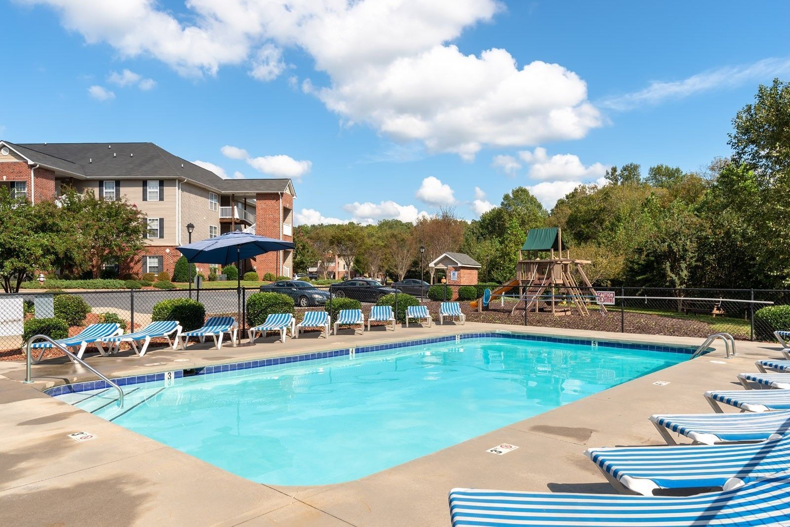 Swimming pool with lounge chairs, apartment building in background, blue sky with clouds.