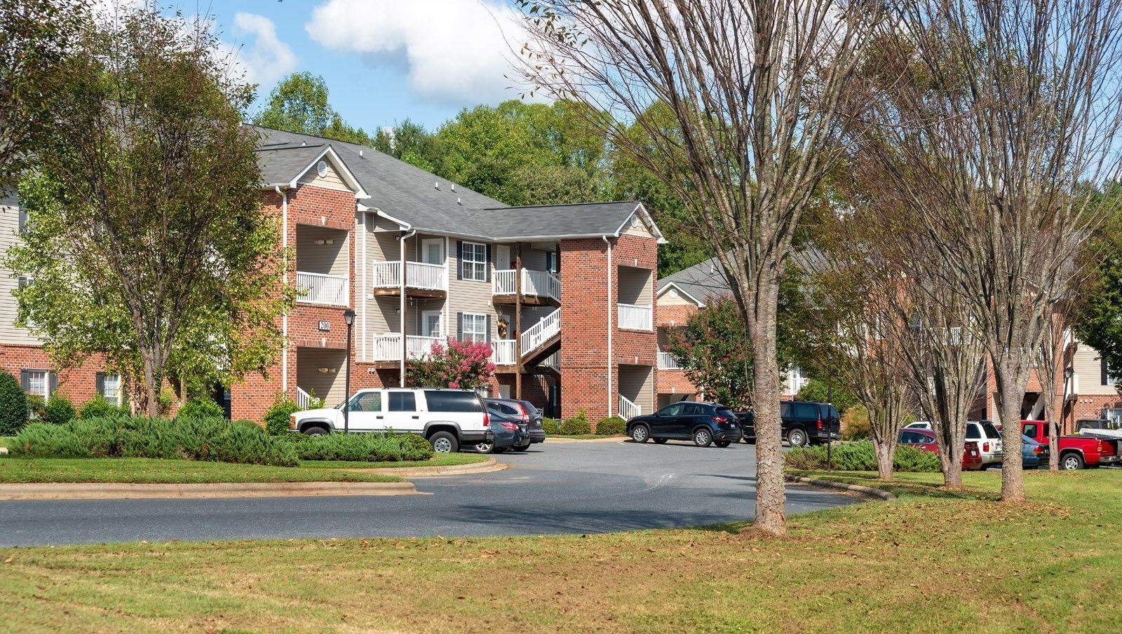 Apartment building exterior, brick and siding, parked cars in front, trees, blue sky.
